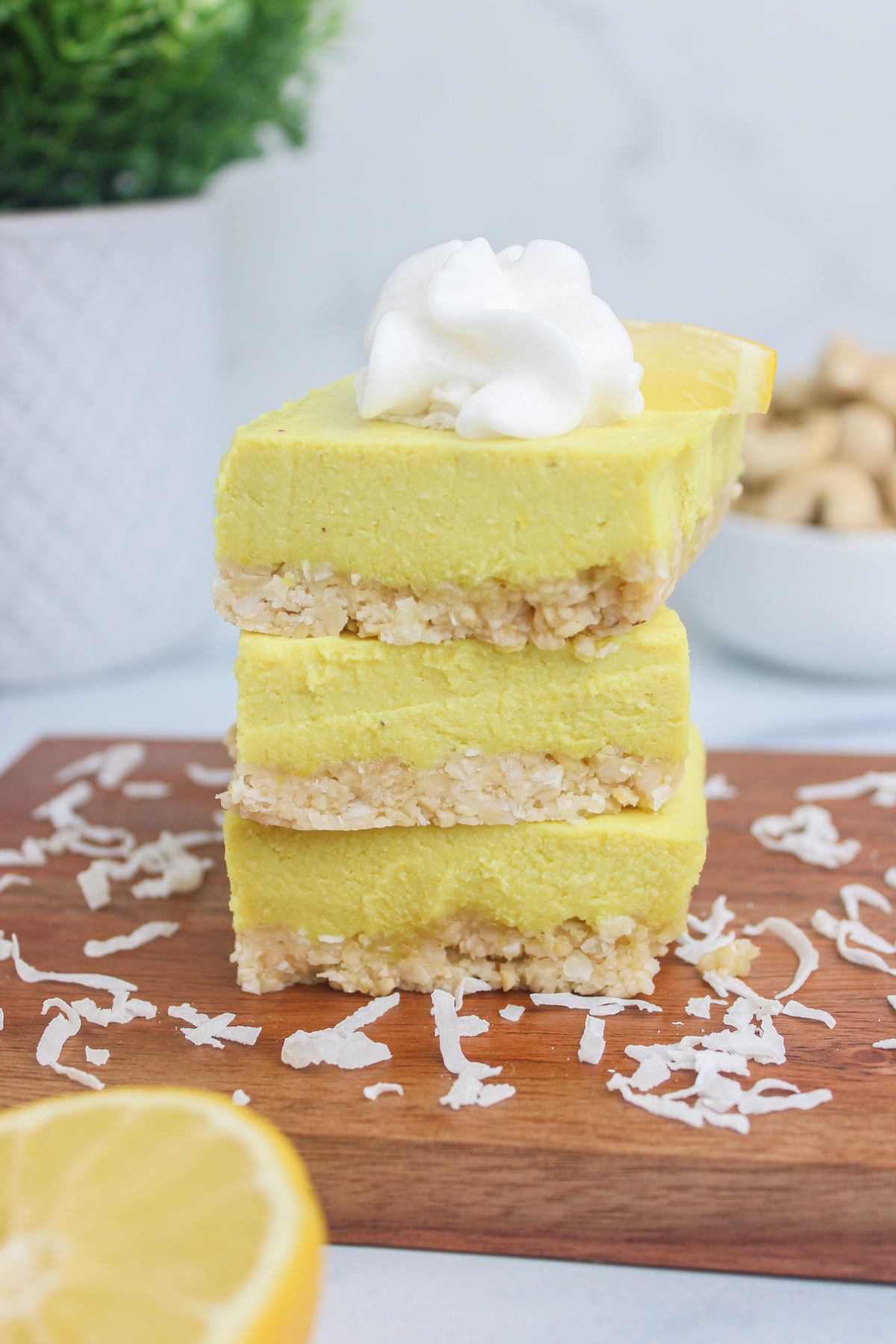 Straight-on shot of three stacked vegan lemon cheesecake bars on a wooden cutting board, showing the creamy lemon filling and crumbly cashew–coconut crust.