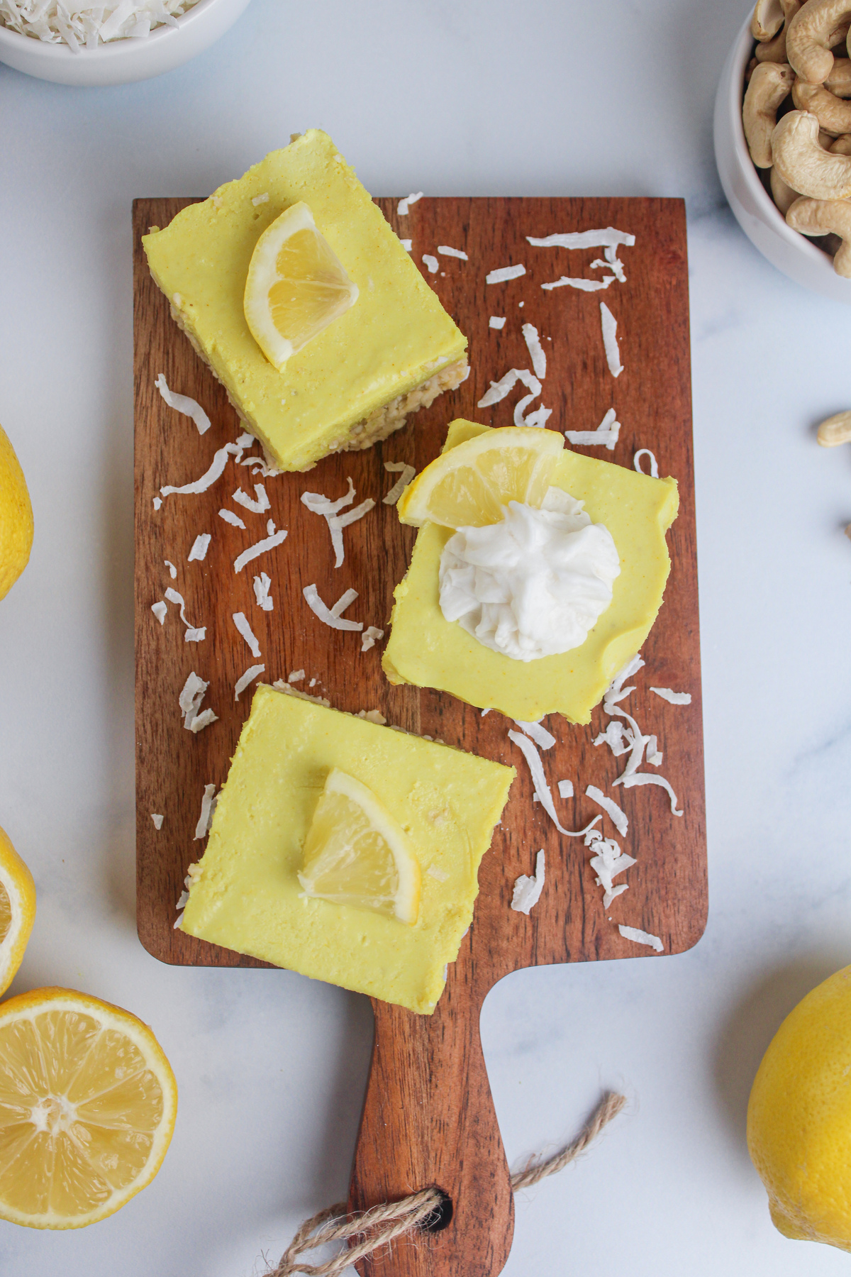 Overhead view of no-bake vegan lemon cheesecake bars arranged on a wooden cutting board, each topped with whipped cream and a small lemon wedge.