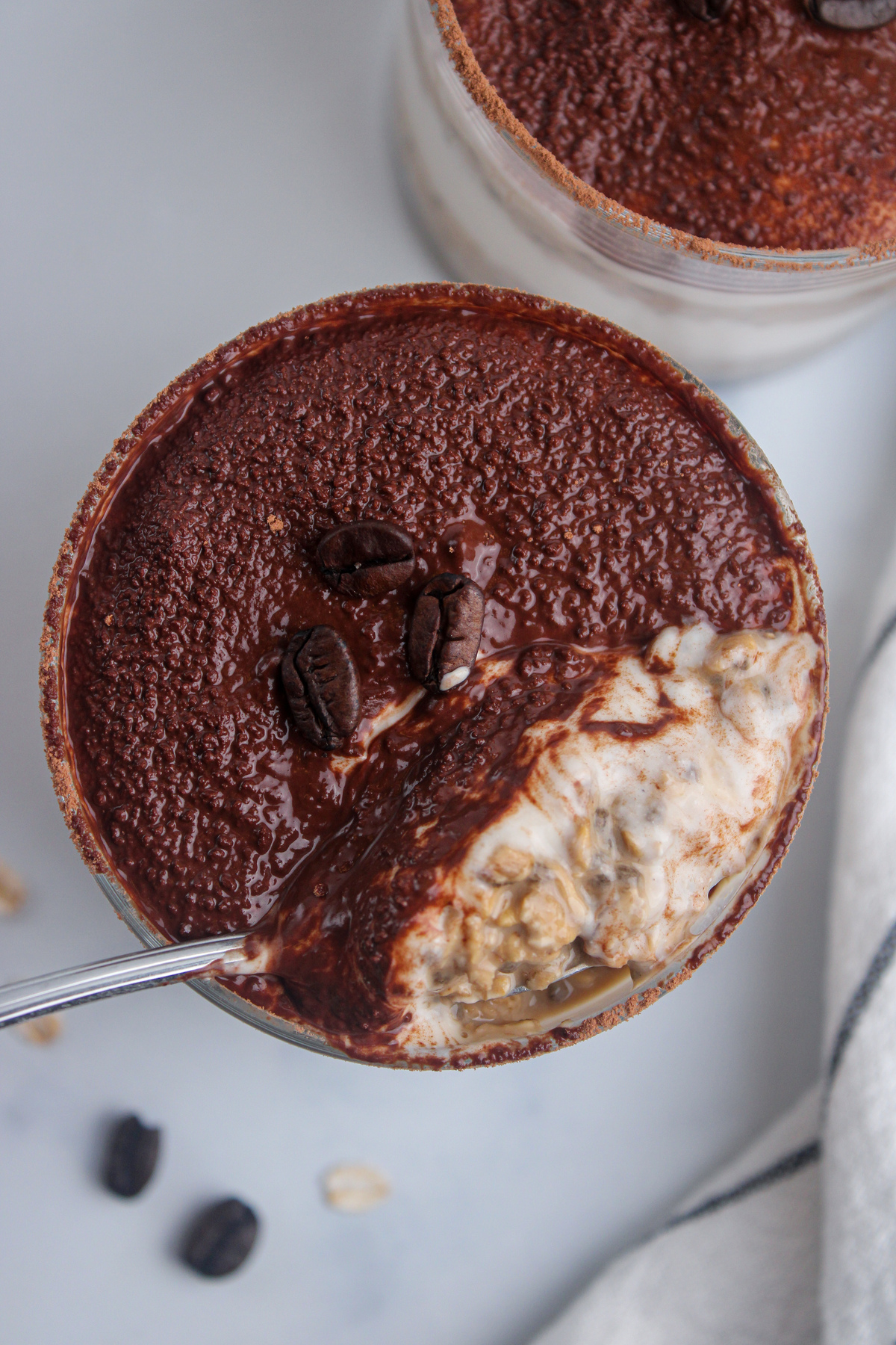 Overhead close-up of tiramisu overnight oats topped with cocoa powder, with a spoon taking a bite.