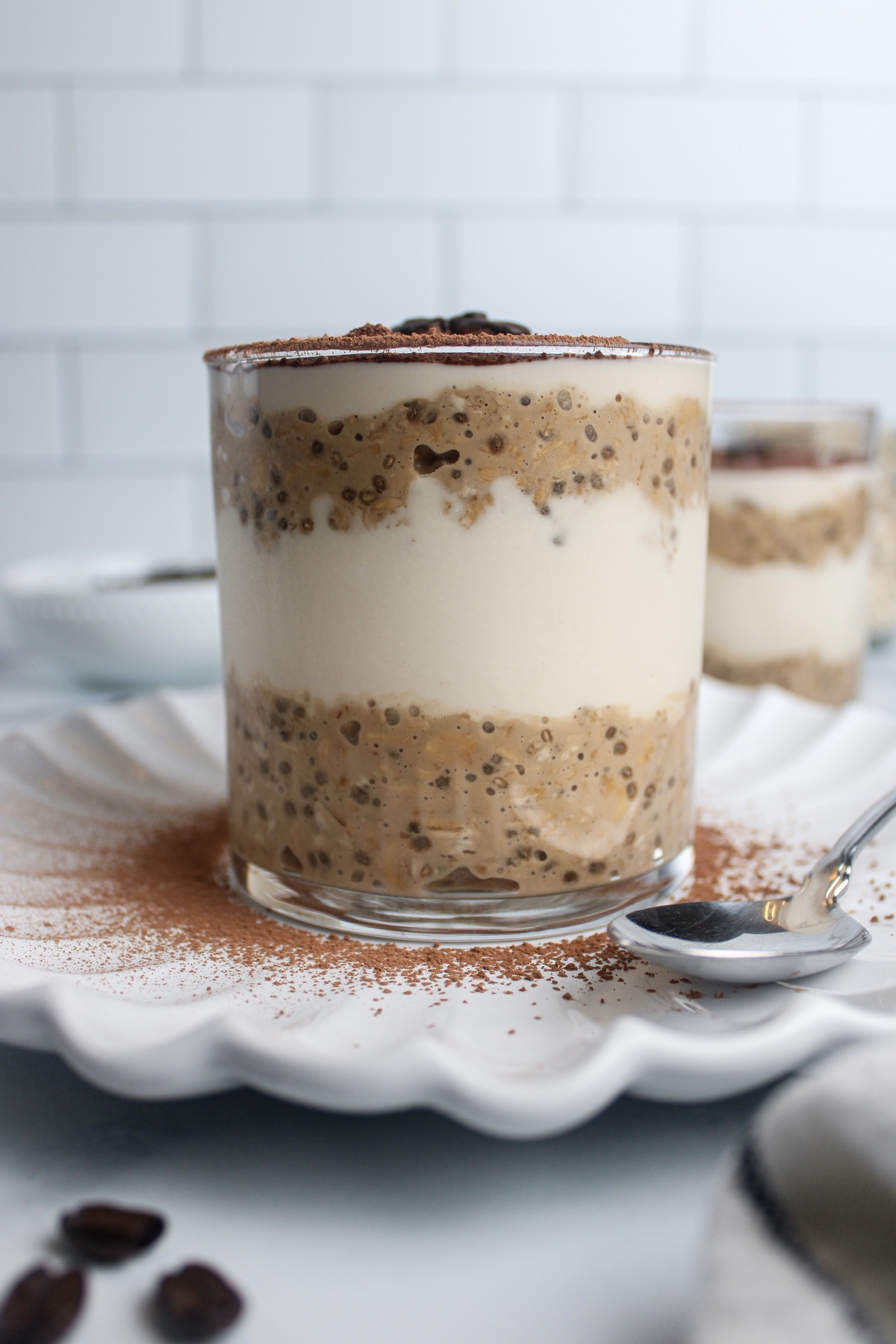 Straight-on view of a glass of tiramisu overnight oats, showing layers of coffee-flavored oats, non-dairy yogurt, and cocoa powder.