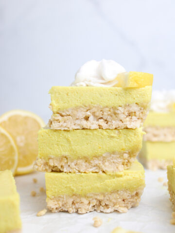 A stack of three no-bake vegan lemon cheesecake bars, with a cashew-coconut crust, topped with whipped cream and a lemon wedge.
