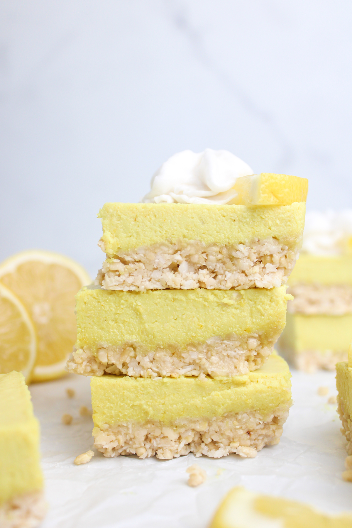 A stack of three no-bake vegan lemon cheesecake bars, with a cashew-coconut crust, topped with whipped cream and a lemon wedge.