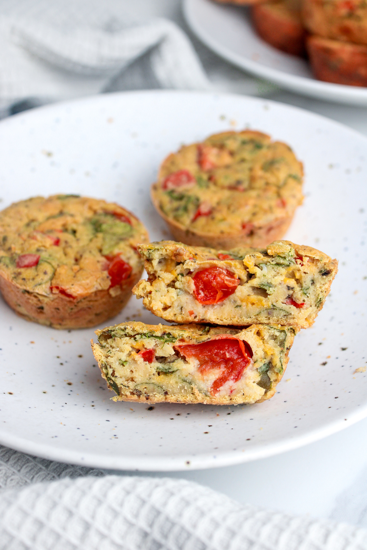 Three savory chickpea flour muffins on a plate, one cut in half to reveal chopped vegetables and vegan cheese inside.