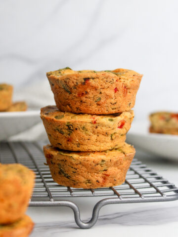 Stack of three savory vegan chickpea flour muffins with vegetables and vegan cheese, arranged on a wire rack.