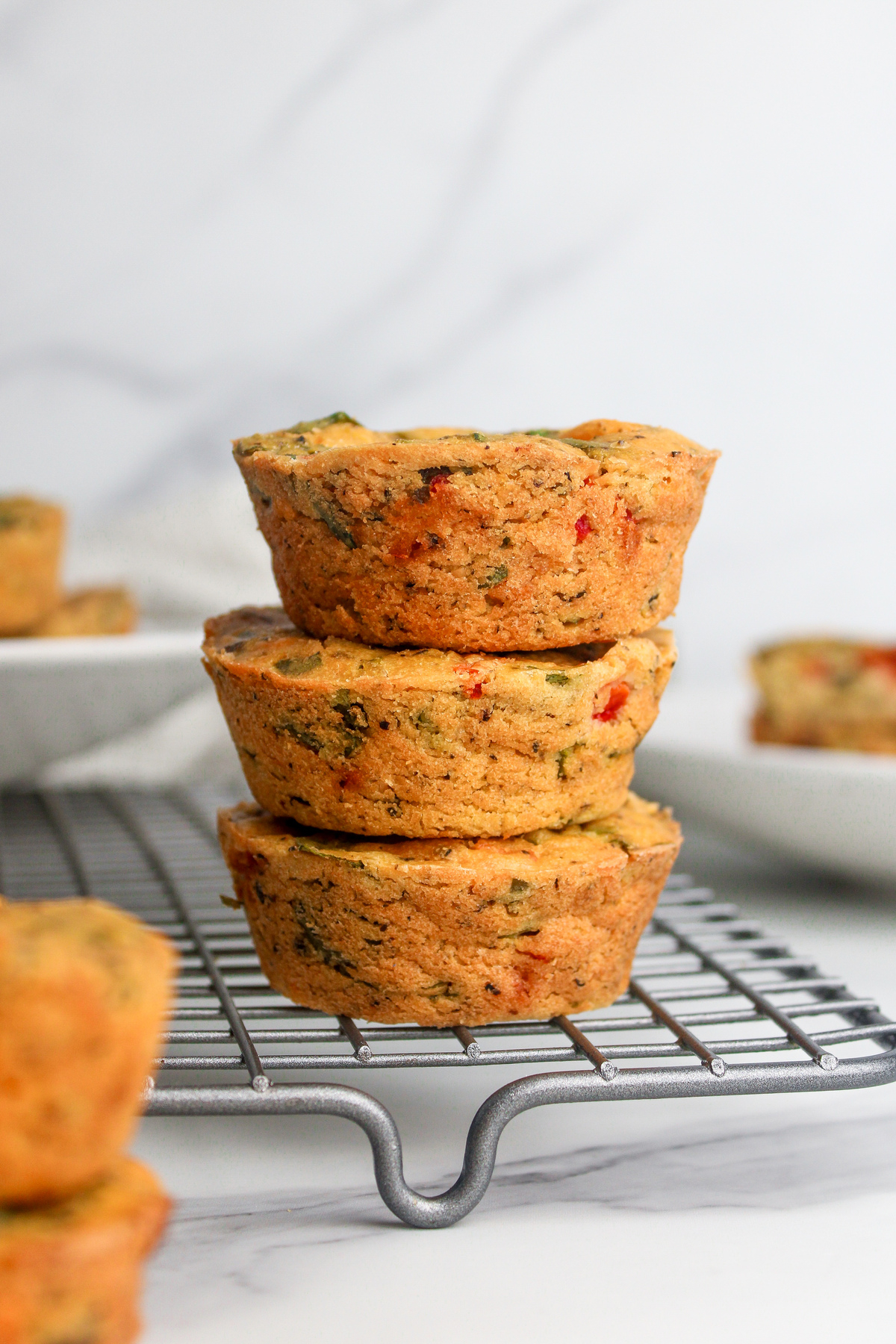 Stack of three savory vegan chickpea flour muffins with vegetables and vegan cheese, arranged on a wire rack.