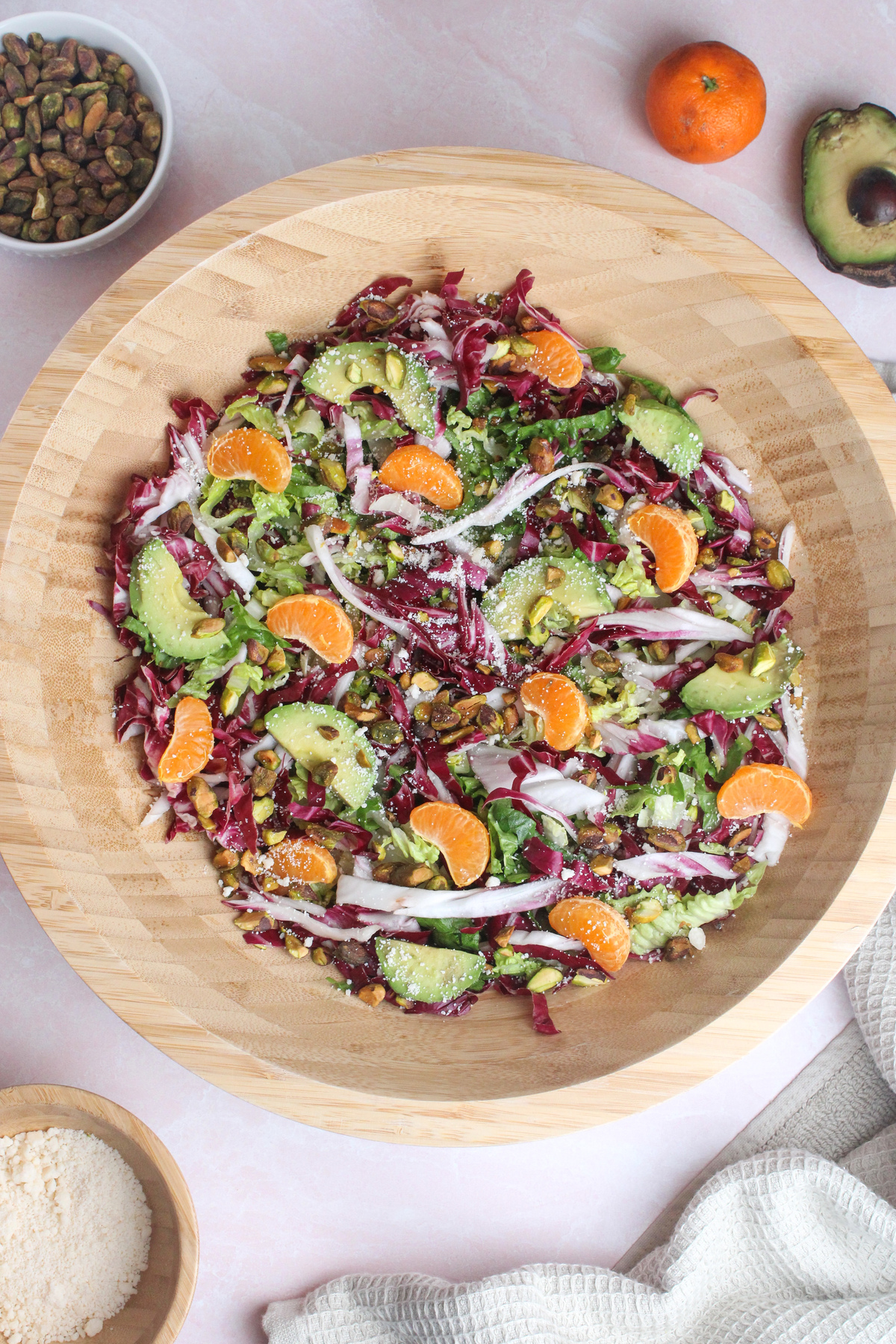 Salad assembled in a large wooden bowl with radicchio, romaine, clementine segments, chopped avocado, pistachios, and vegan parmesan.