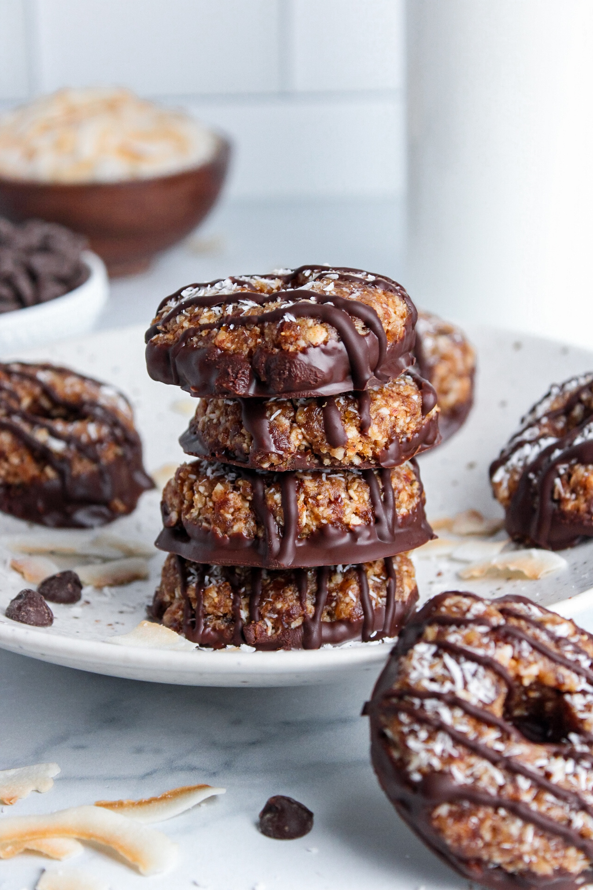 Stack of no-bake Samoa cookies on a plate, with additional cookies, toasted coconut, and chocolate chips scattered around.