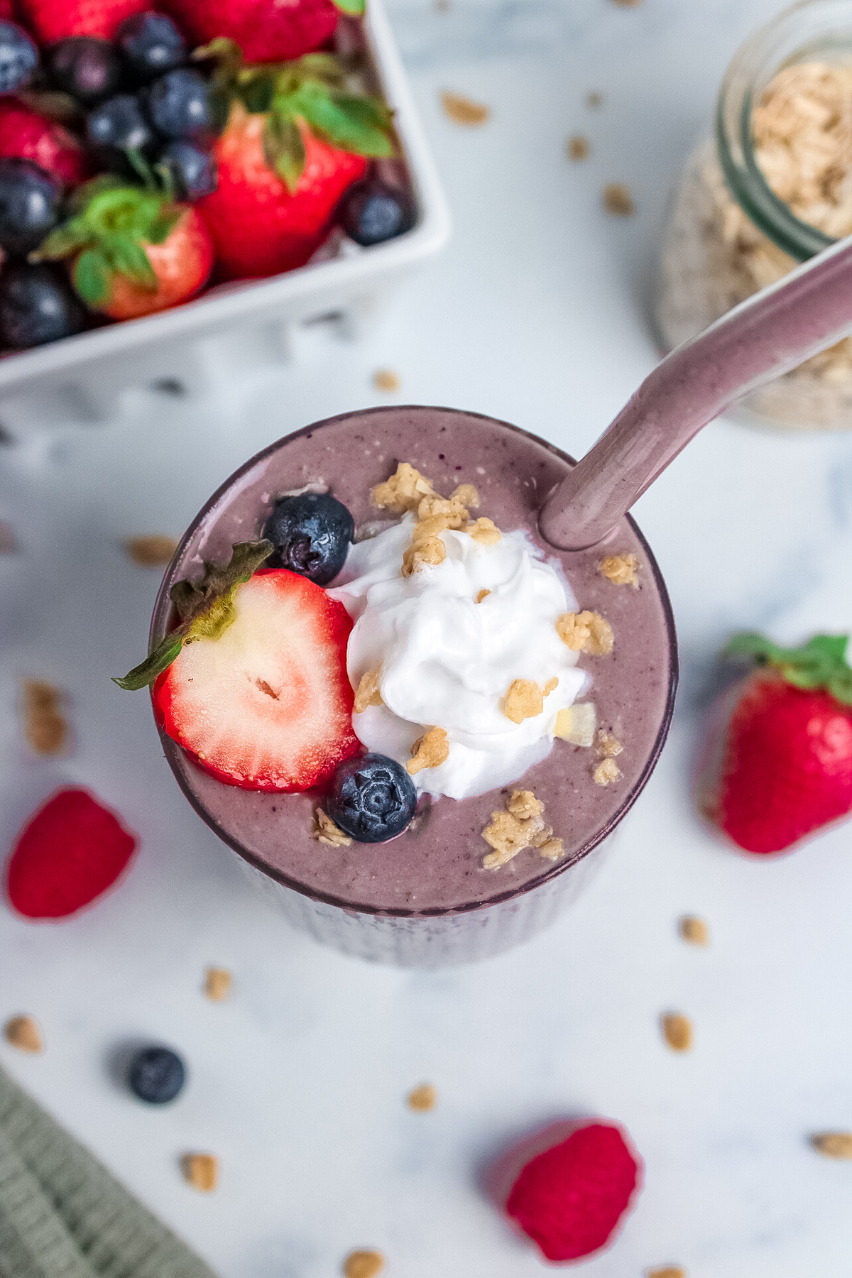 Overhead view of a high-protein berry pie smoothie topped with fresh berries, whipped cream, and granola, with a straw.