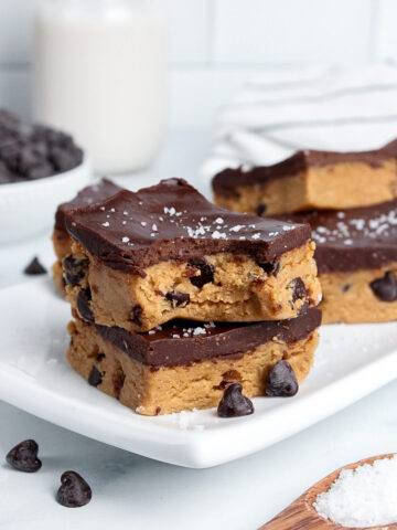 Stack of two healthy no-bake cookie dough bars with chocolate chips inside and chocolate ganache on top, one with a bite taken out.