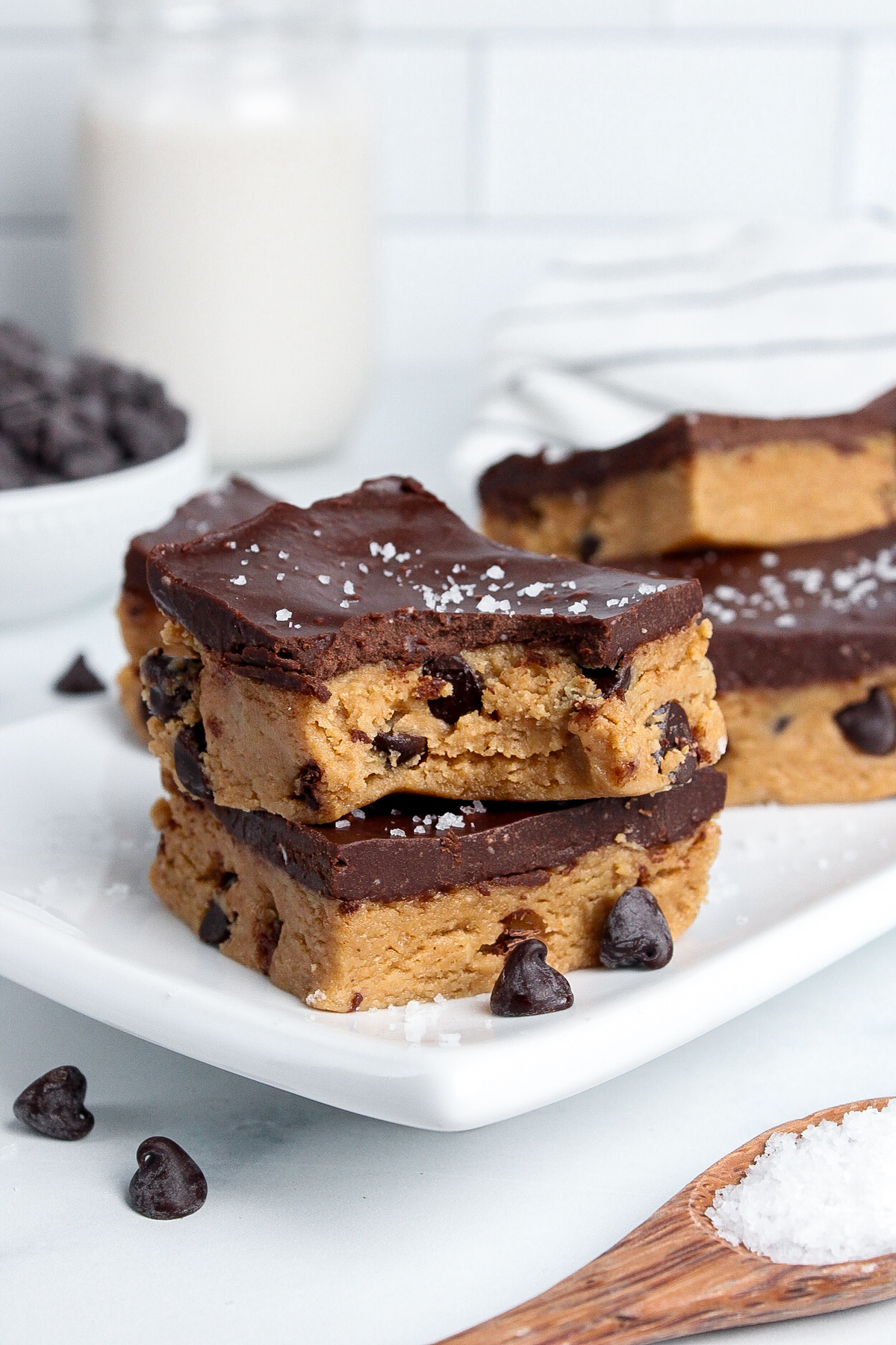 Stack of two healthy no-bake cookie dough bars with chocolate chips inside and chocolate ganache on top, one with a bite taken out.