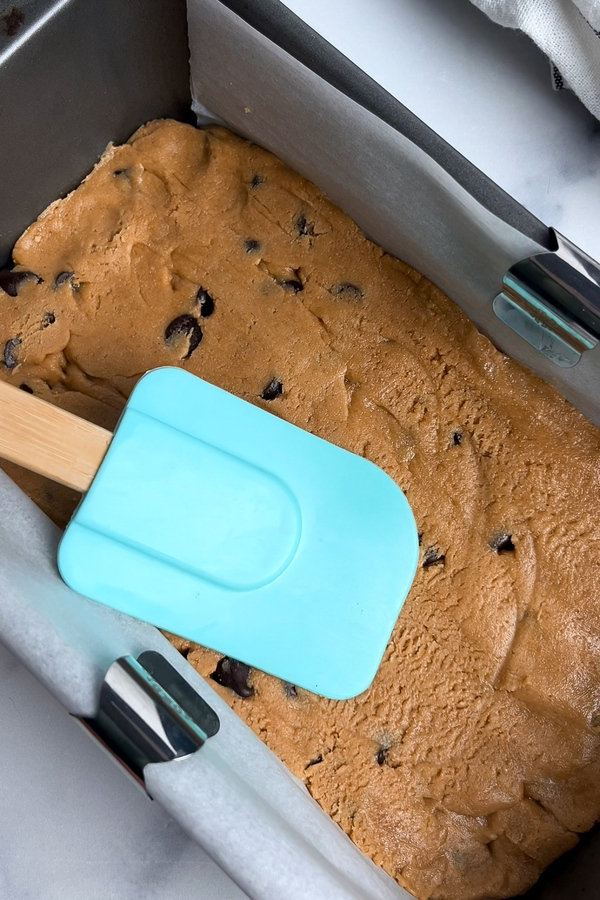 Pressing no-bake cookie dough into a parchment-lined pan using a rubber spatula.