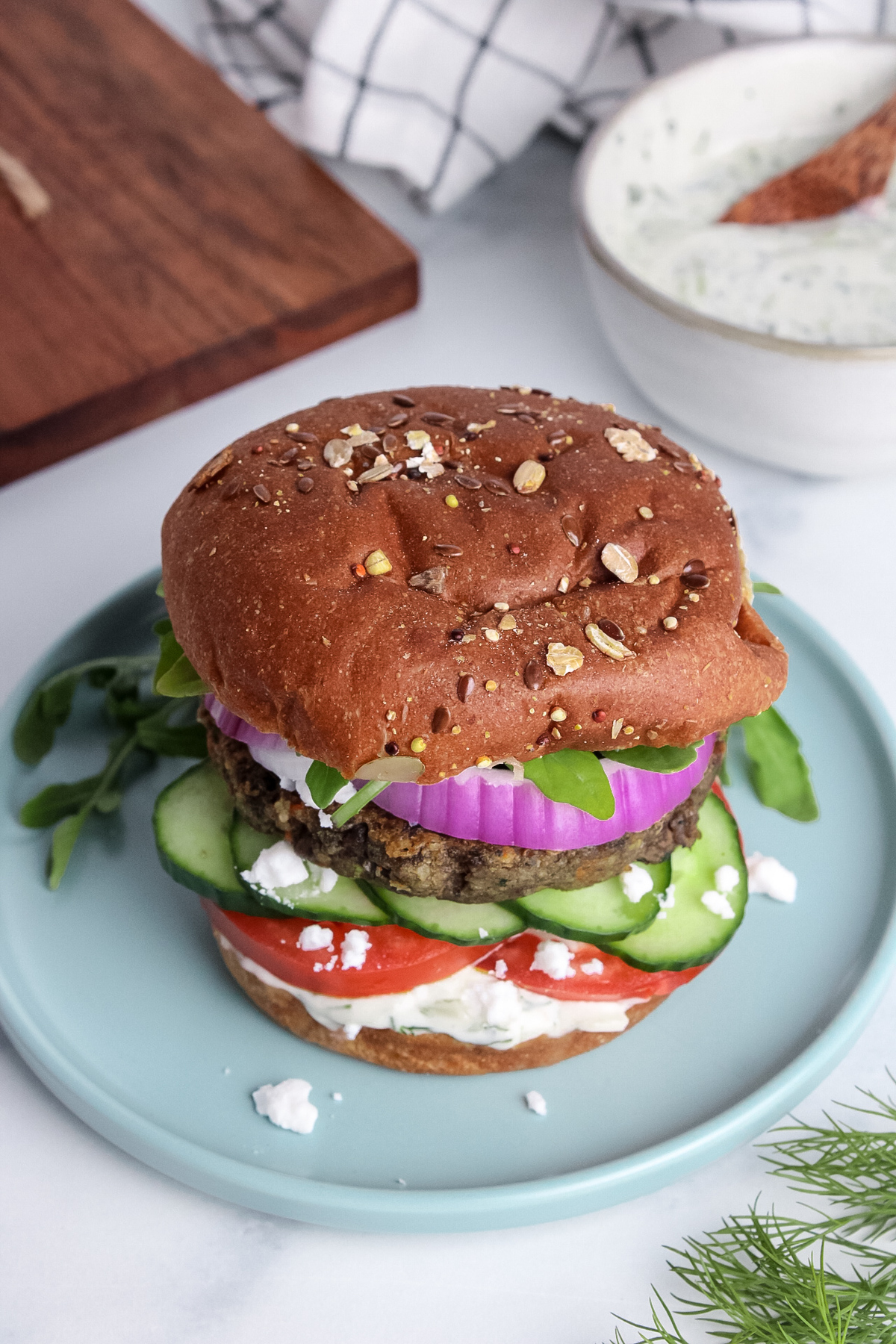 Angled view of a Mediterranean veggie burger on a toasted whole-grain bun, piled high with fresh vegetables and dairy-free tzatziki.