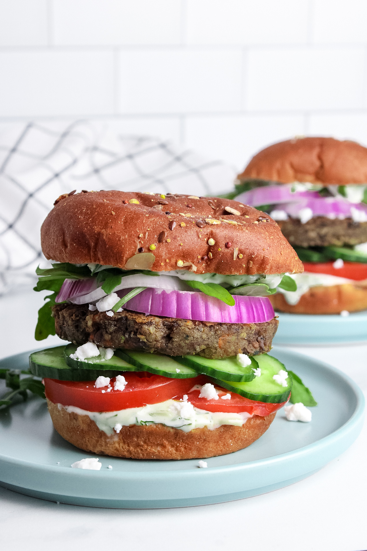 Two Mediterranean veggie burgers on plates, stacked in toasted buns with colorful vegetables and creamy tzatziki sauce.