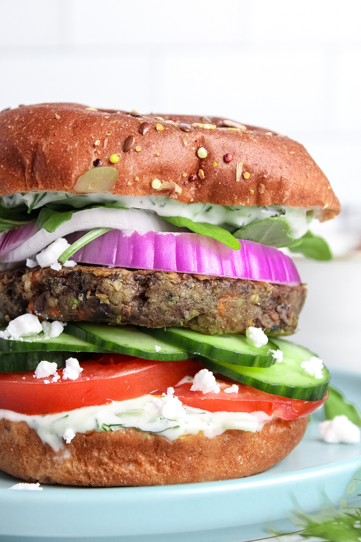 Close-up of a Mediterranean-themed burger with a plant-based patty, vegan feta, tzatziki, tomato, cucumber, red onion, and arugula.