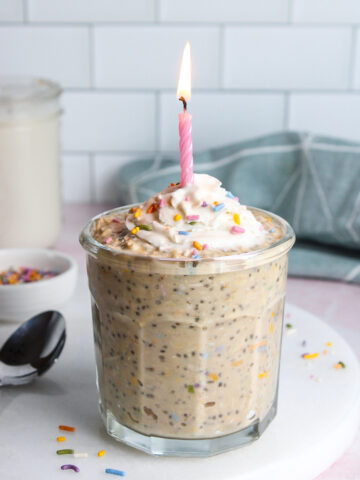Birthday cake overnight oats in a glass, topped with dairy-free whipped cream, colorful sprinkles, and a lit birthday candle.