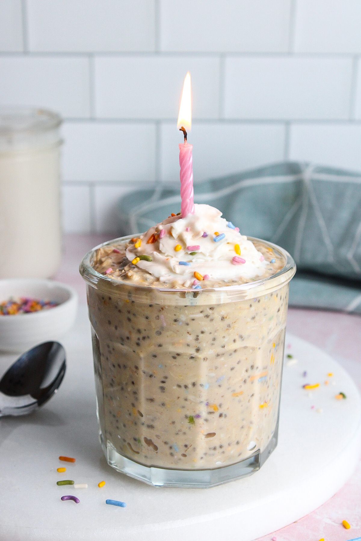 Birthday cake overnight oats in a glass, topped with dairy-free whipped cream, colorful sprinkles, and a lit birthday candle.