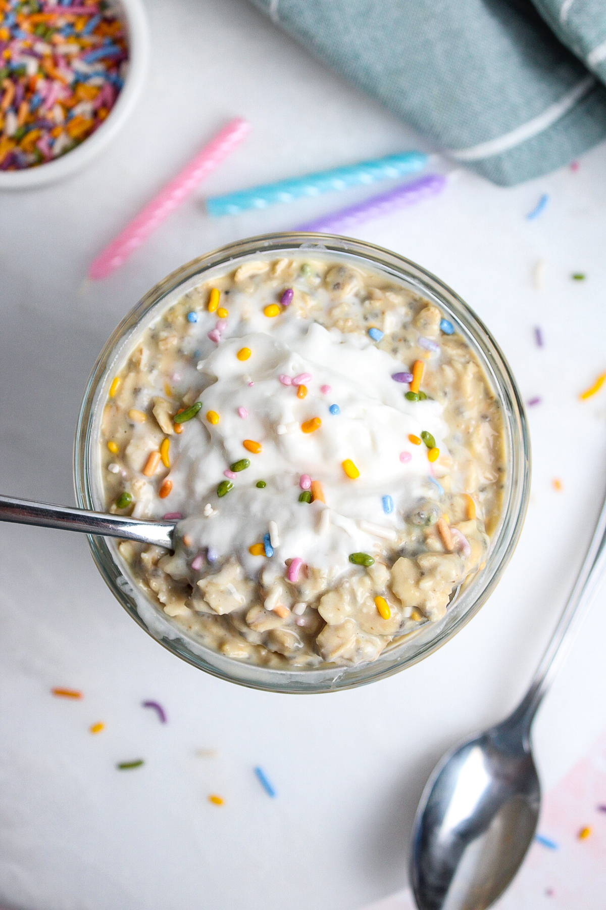Overhead close-up of birthday cake overnight oats in a glass, with a spoon scooping into the thick, creamy mixture.