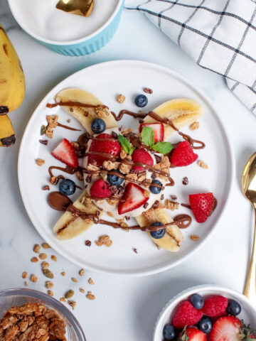 Overhead view of a breakfast banana split on a plate, with a halved banana topped with yogurt, granola, fresh berries, and a drizzle of chocolate spread.