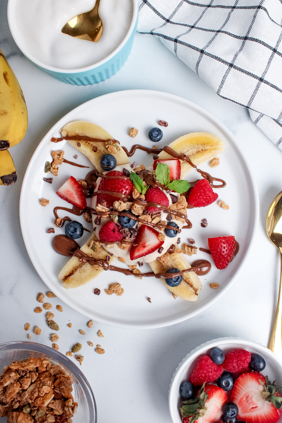 Overhead view of a breakfast banana split on a plate, with a halved banana topped with yogurt, granola, fresh berries, and a drizzle of chocolate spread.