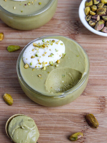 Vibrant green, creamy vegan pistachio mousse in a small dessert glass, topped with whipped coconut cream and crushed pistachios, with a bite taken out.