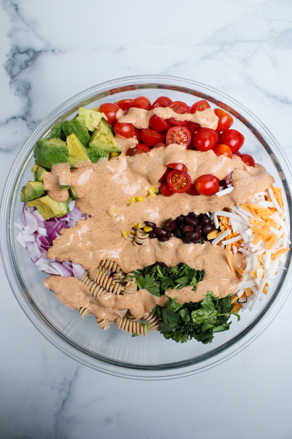 Cooked rotini pasta, black beans, corn, tomatoes, avocado, red onion, cilantro, and shredded non-dairy cheddar cheese added to a large bowl, with the taco-seasoned dressing drizzled on top.