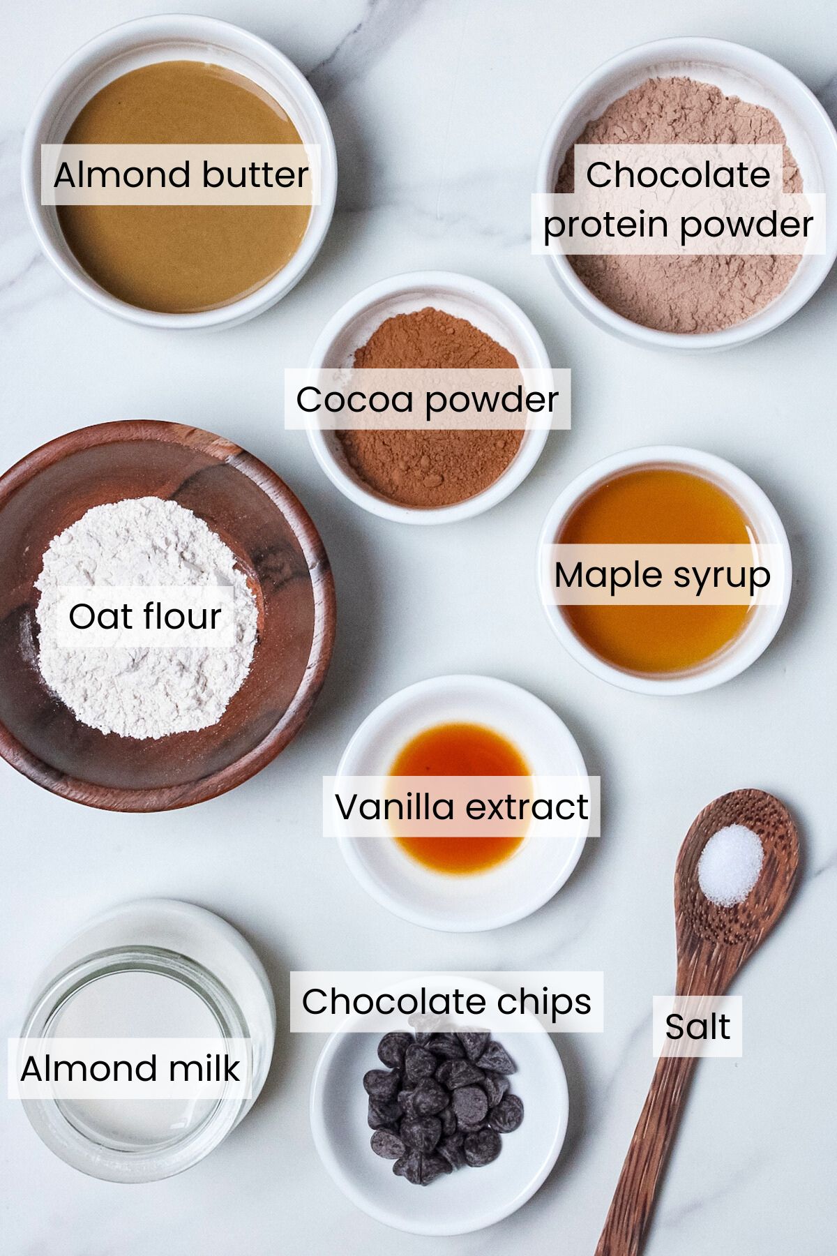 Ingredients for single-serve protein brownie batter: almond butter, chocolate protein powder, cocoa powder, oat flour, maple syrup, vanilla extract, almond milk, chocolate chips, and salt.