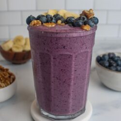 A vibrant purple blueberry banana walnut smoothie in a glass, topped with banana slices, blueberries, and walnuts.