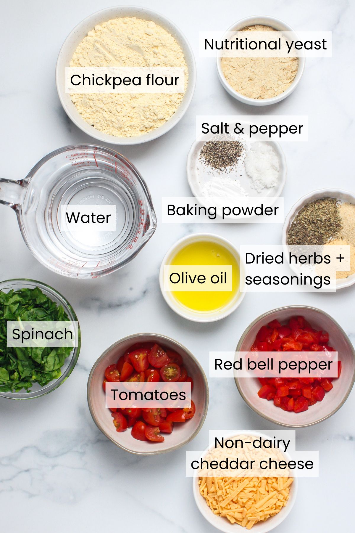 Ingredients needed to make savory vegan chickpea flour muffins: chickpea flour, nutritional yeast, water, salt, pepper, baking powder, olive oil, dried herbs and seasonings, spinach, tomatoes, red bell pepper, and non-dairy cheddar cheese.