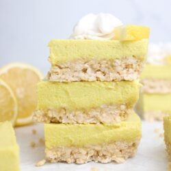 A stack of three no-bake vegan lemon cheesecake bars, with a cashew-coconut crust, topped with whipped cream and a lemon wedge.