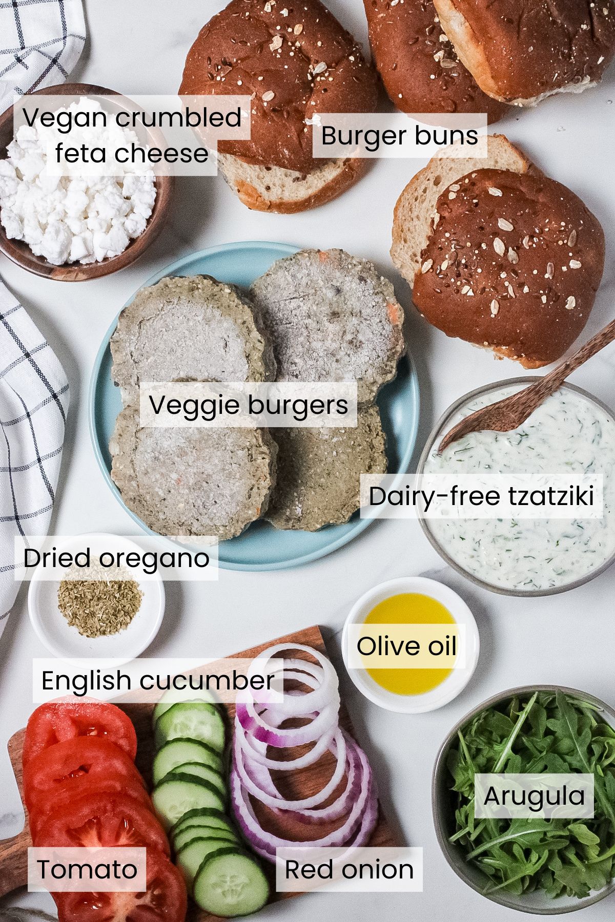 Ingredients for Mediterranean veggie burgers: olive oil, veggie burger patties, dried oregano, burger buns, vegan crumbled feta, tomato, English cucumber, red onion, arugula, and homemade dairy-free tzatziki.