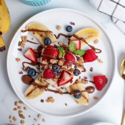 Overhead view of a breakfast banana split on a plate, with a halved banana topped with yogurt, granola, fresh berries, and a drizzle of chocolate spread.