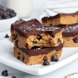 Stack of two healthy no-bake cookie dough bars with chocolate chips inside and chocolate ganache on top, one with a bite taken out.