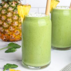 Two glasses of pineapple protein green smoothie with wooden straws, garnished with shredded coconut and pineapple wedges.