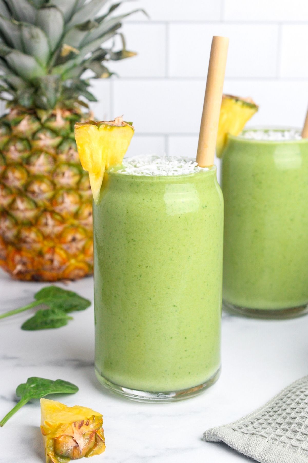 Two glasses of pineapple protein green smoothie with wooden straws, garnished with shredded coconut and pineapple wedges.