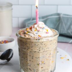 Birthday cake overnight oats in a glass, topped with dairy-free whipped cream, colorful sprinkles, and a lit birthday candle.