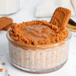 Creamy Biscoff overnight oats in a glass jar with a cookie butter layer, crushed Biscoff cookies around the rim, and a whole cookie on top.