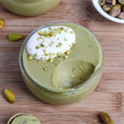 Vibrant green, creamy vegan pistachio mousse in a small dessert glass, topped with whipped coconut cream and crushed pistachios, with a bite taken out.