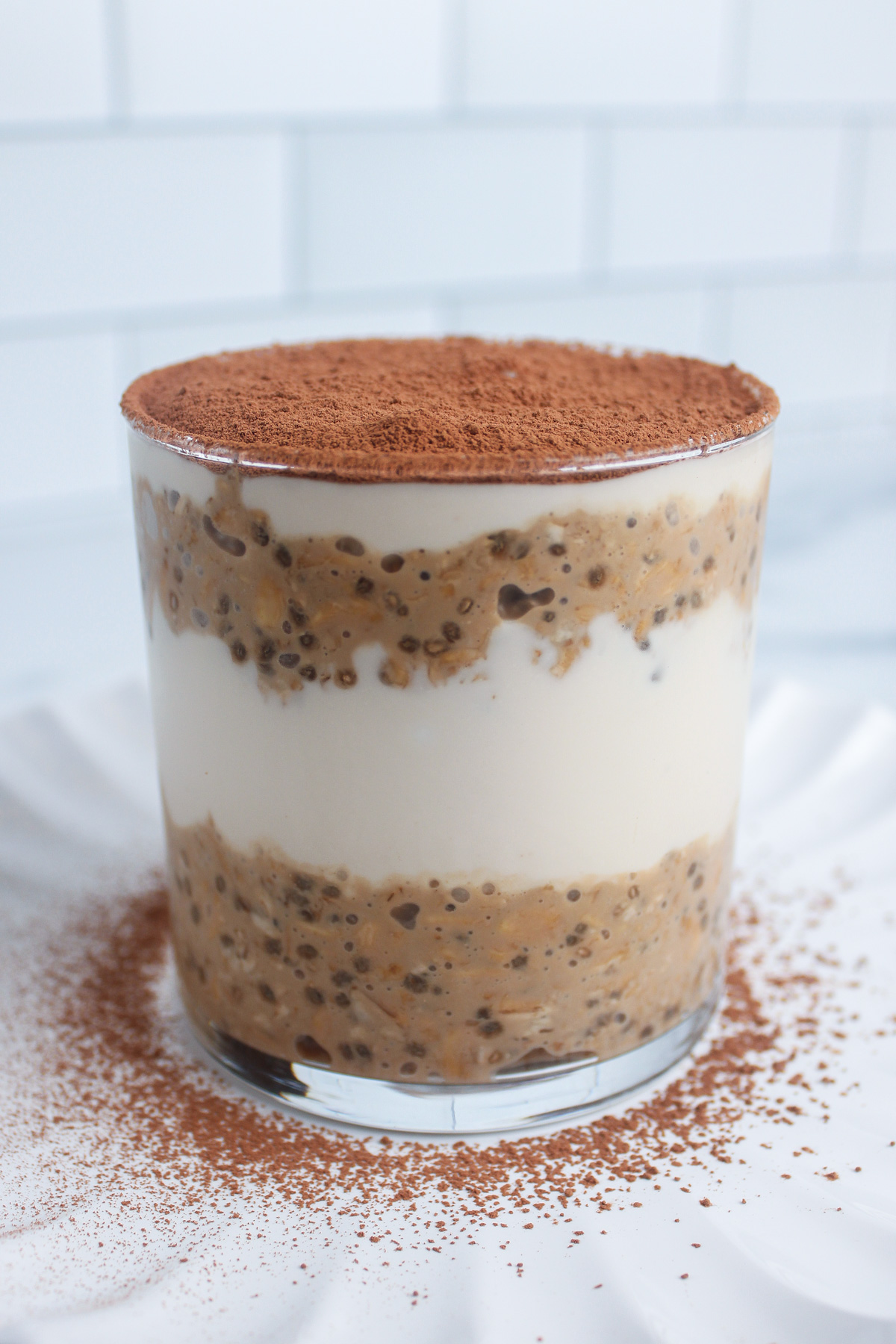 Alternating layers of coffee overnight oats and non-dairy yogurt in a glass, topped with a dusting of cocoa powder.